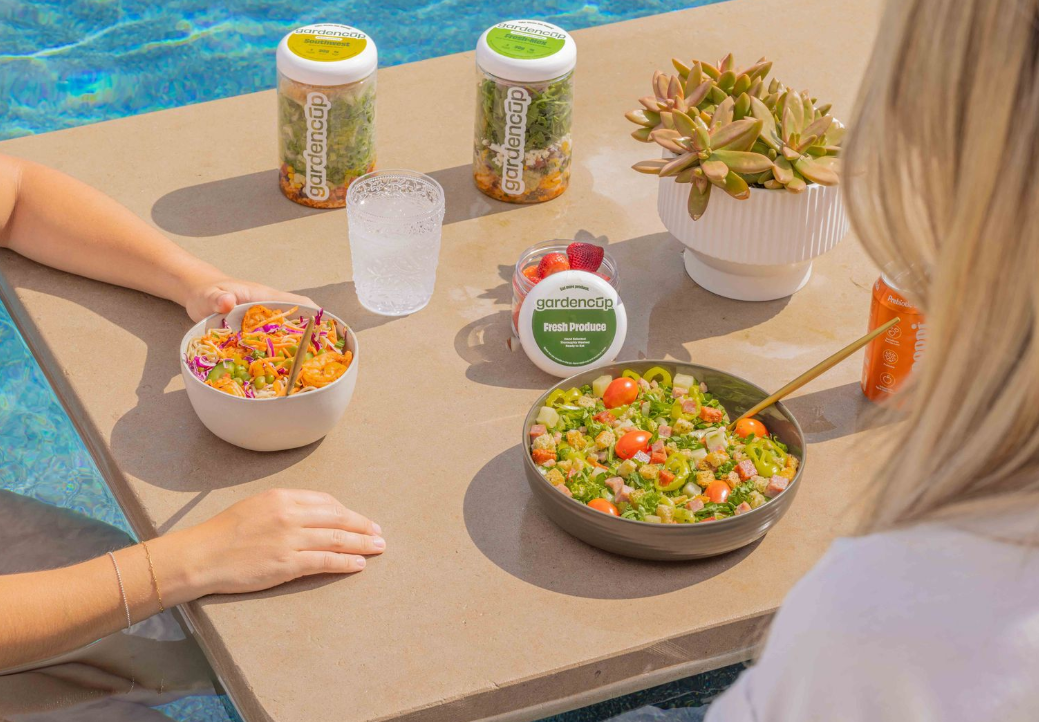 Two people eating salads by a pool with garden cups and a plant.