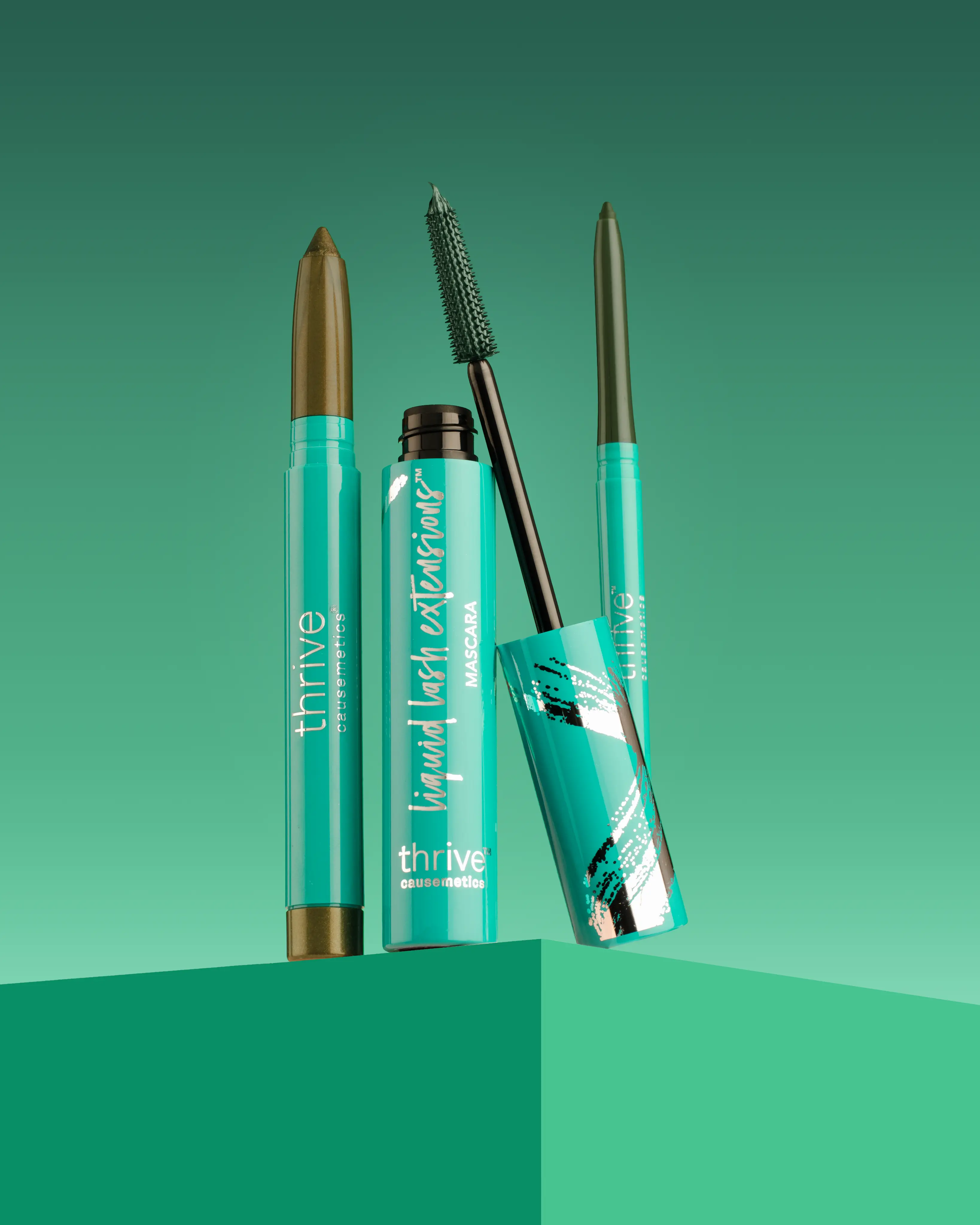 A collection of Thrive Causemetics mascara, eyeliner, and eyeshadow stick on a green, geometric background.