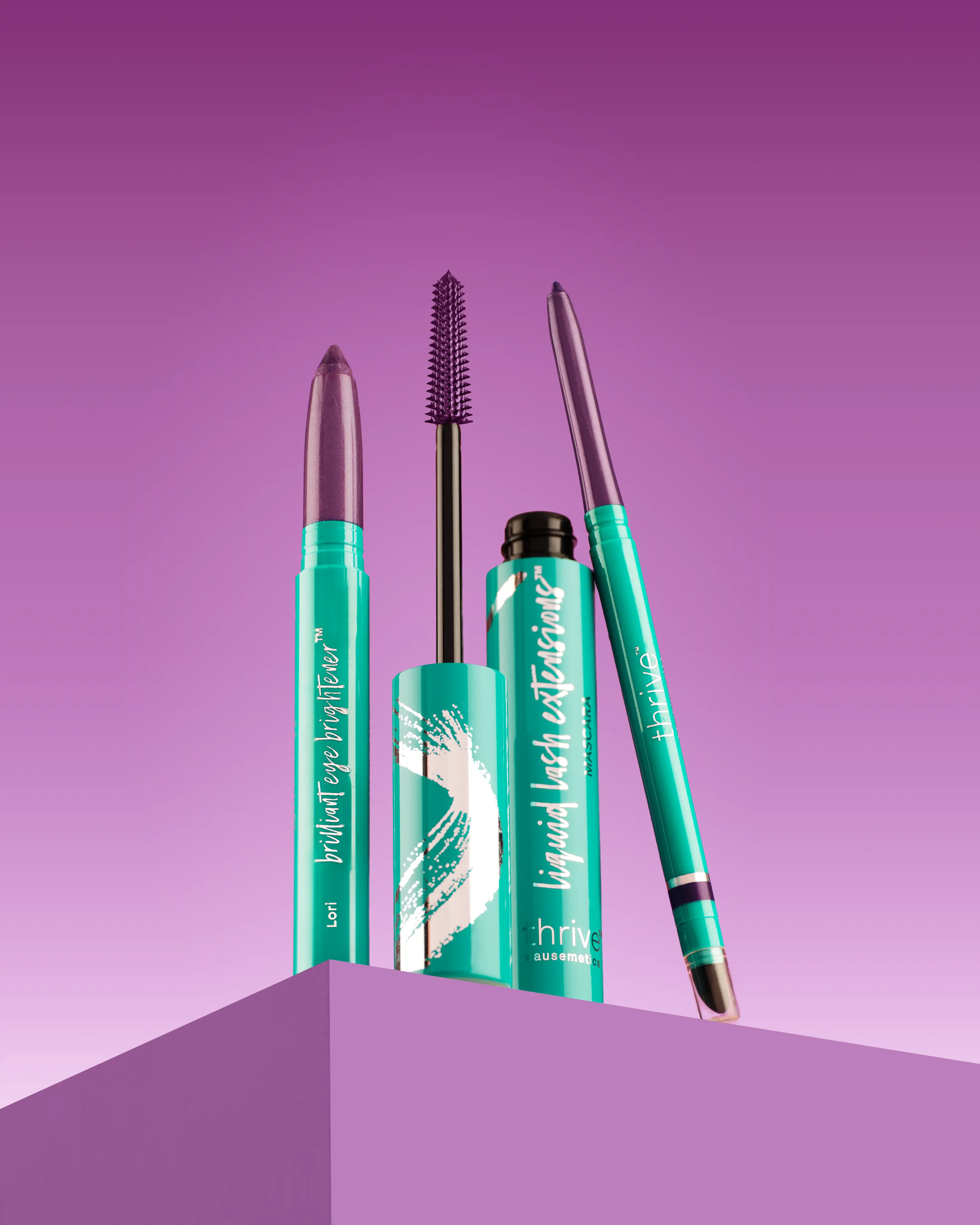 Three eye makeup products with teal packaging and purple accents against a purple background.