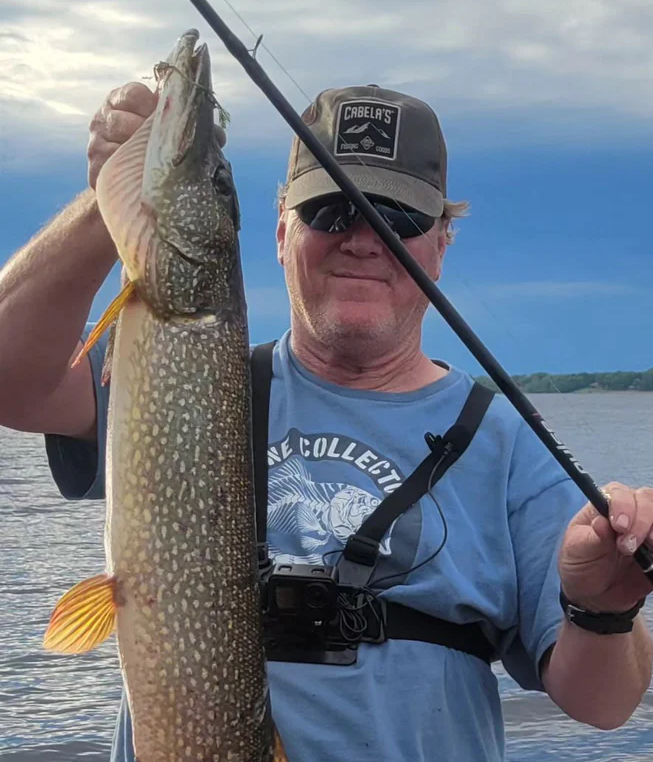 Person holding a large fish and fishing rod by a lake.