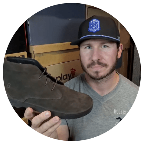 Person holding a brown shoe, wearing a cap and gray t-shirt, with boxes in the background.