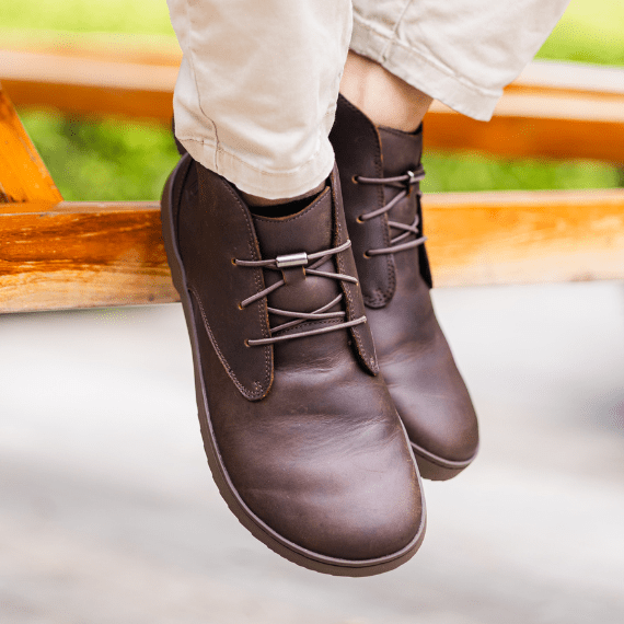 Person wearing brown leather shoes and beige pants sitting on a wooden bench.