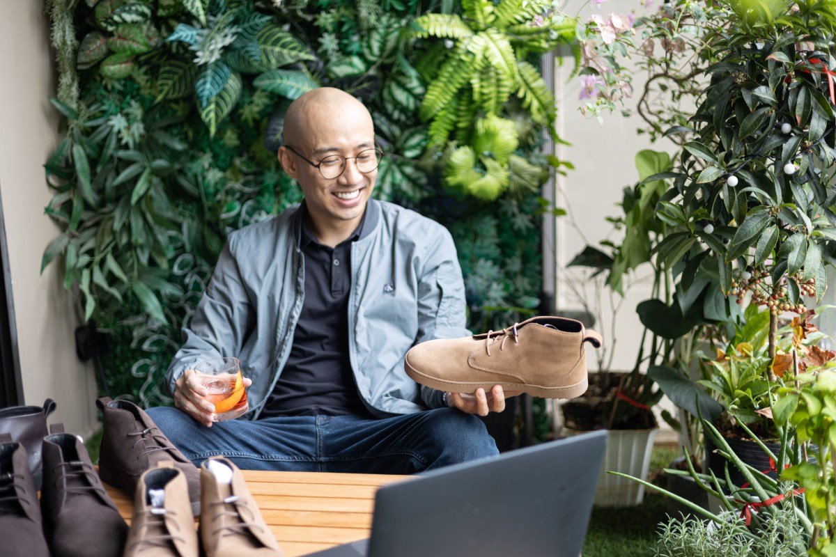 Person holding a shoe and smiling in a garden setting, with a laptop and drink nearby.