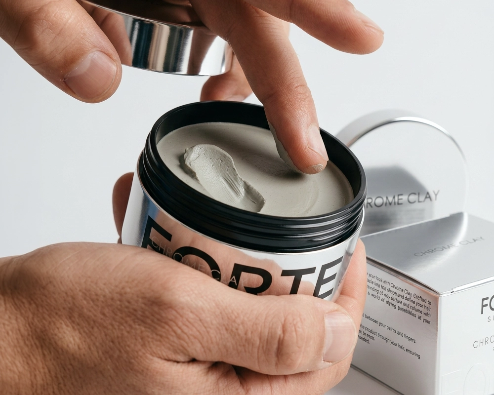A person dips their finger into an open jar of Forte Chrome Clay hair styling product.
