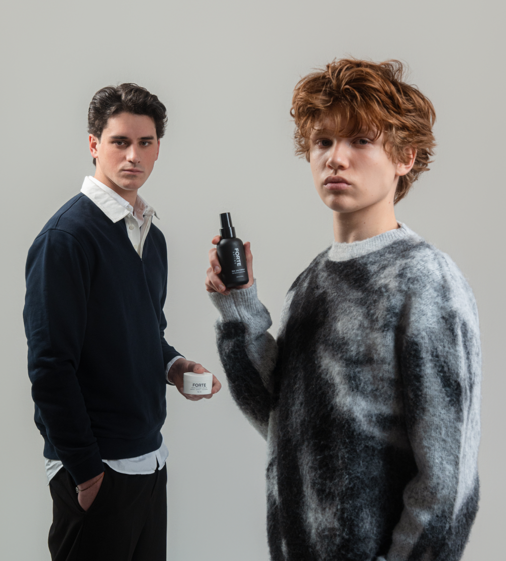 Two young men hold up Forte brand hair products, one a white jar and one a black spray bottle.