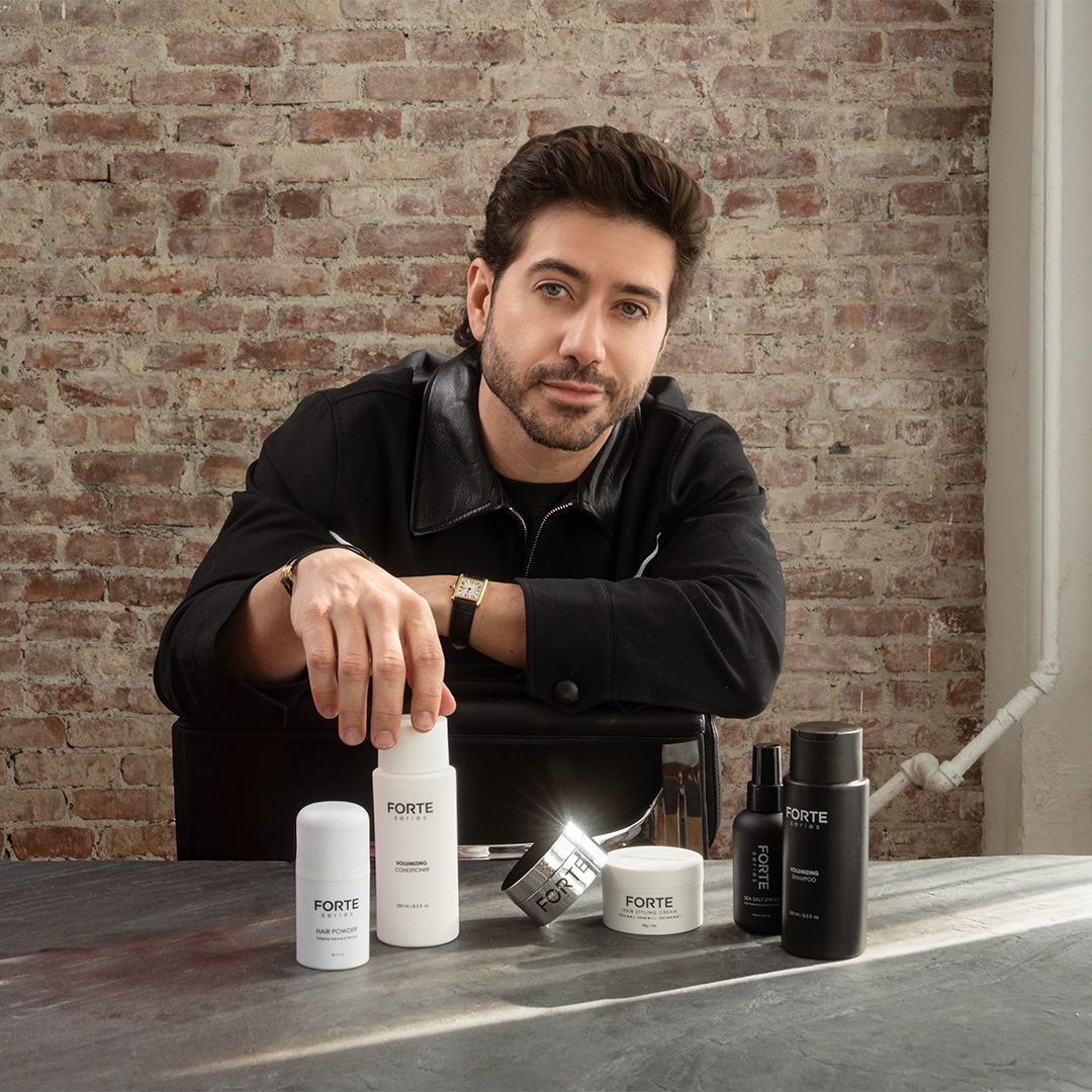 A man in a black jacket sits at a table with several Forte Series hair care products.
