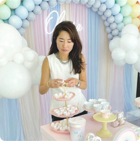 Woman arranging dessert stand at a pastel-themed event with balloon arch.