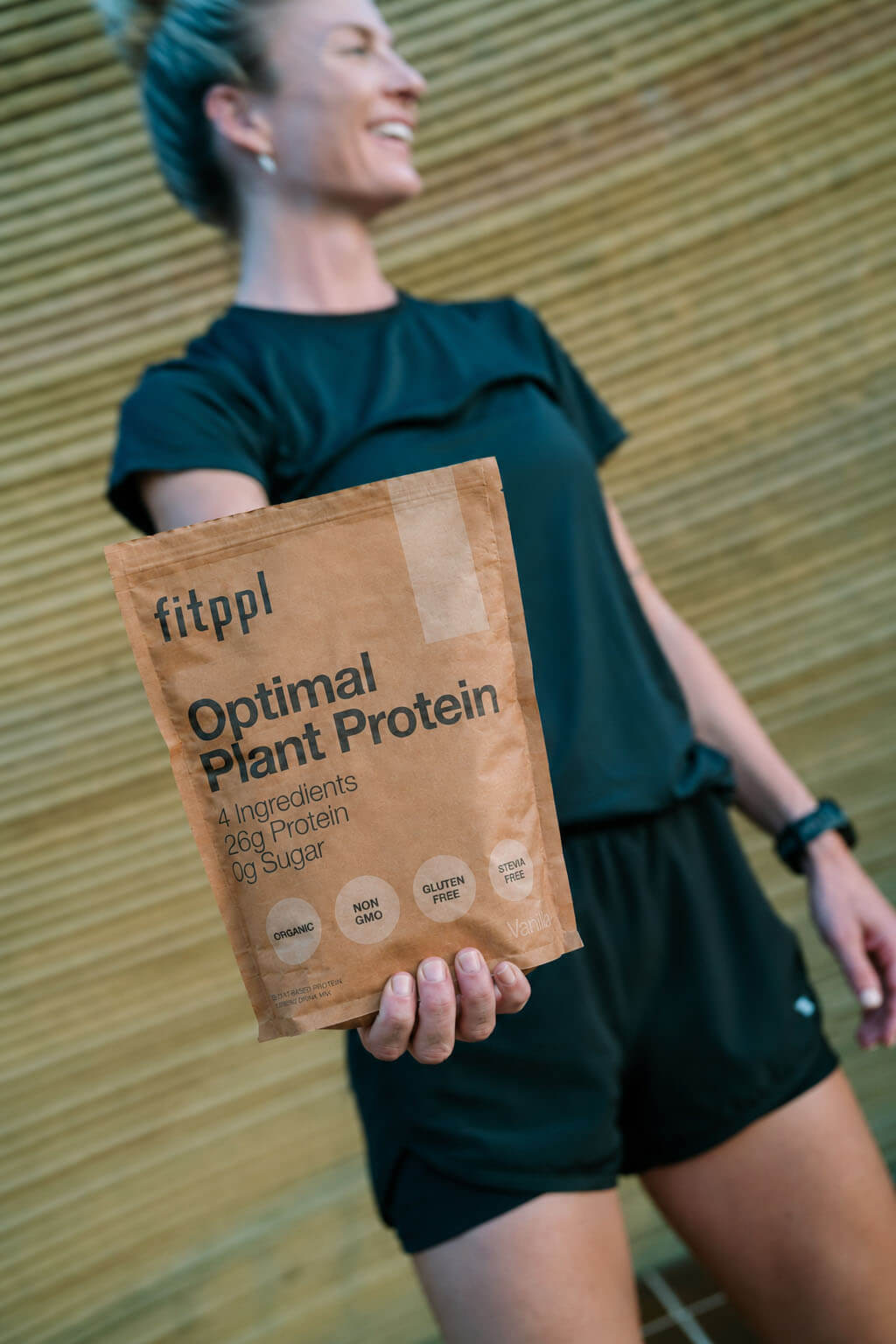 A smiling woman in athletic clothes holds a bag of Fitppl Optimal Plant Protein.