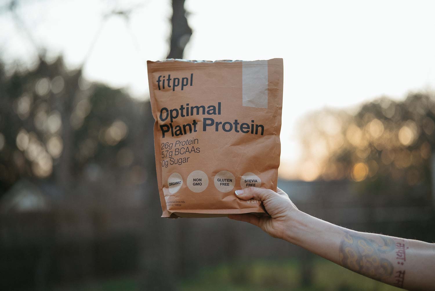 A person's tattooed arm holds a bag of fitppl Optimal Plant Protein powder outdoors.