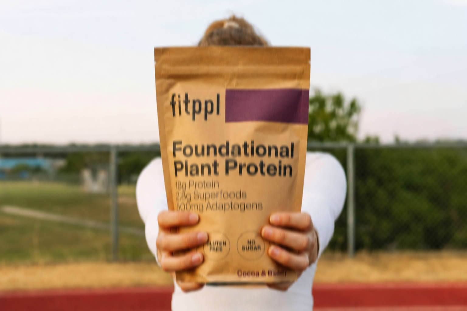 A person in a white shirt holds a bag of fitppl Foundational Plant Protein outdoors on a track.