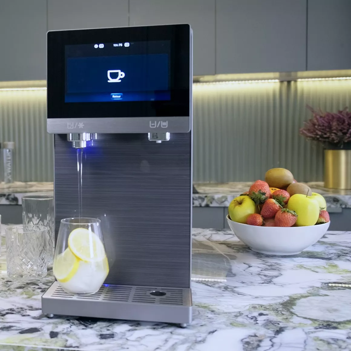 A sleek water dispenser pours water into a glass with lemon and ice on a marble kitchen counter.
