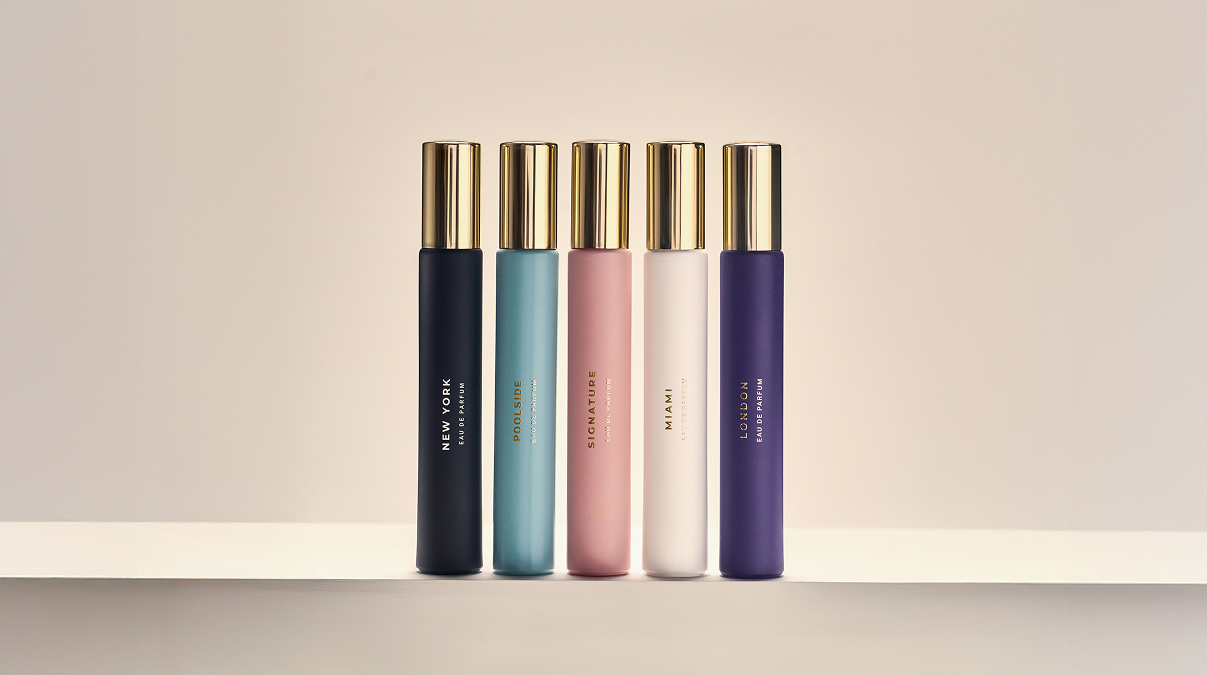 Six different colored travel-size perfume bottles with gold caps lined up on a white shelf.