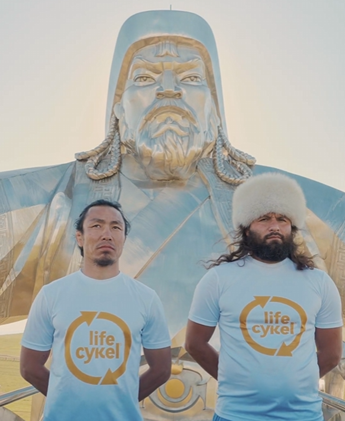 Two men in blue shirts stand before a large metallic statue.