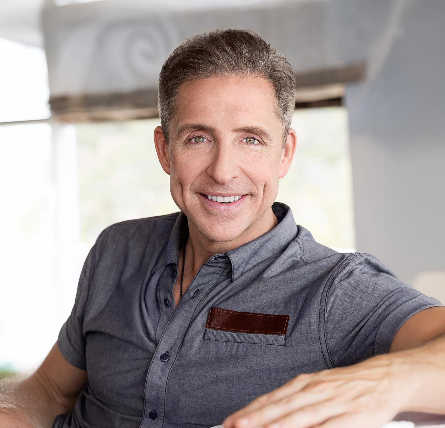 Smiling man in a gray shirt, sitting in a bright room.