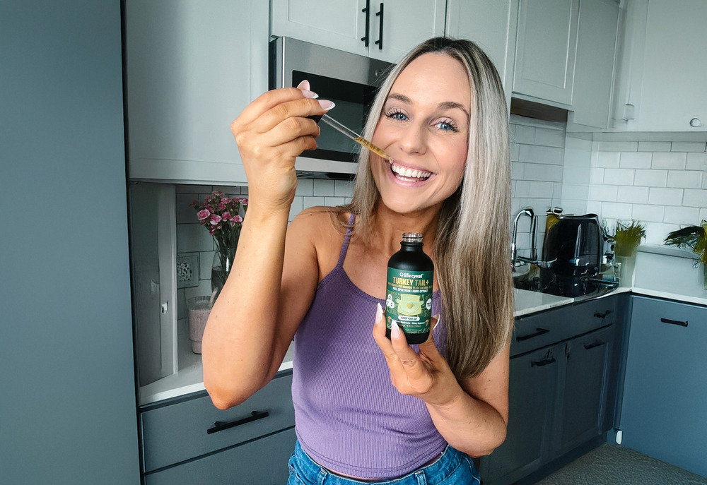 Woman in kitchen holding a bottle and dropper, smiling at the camera.