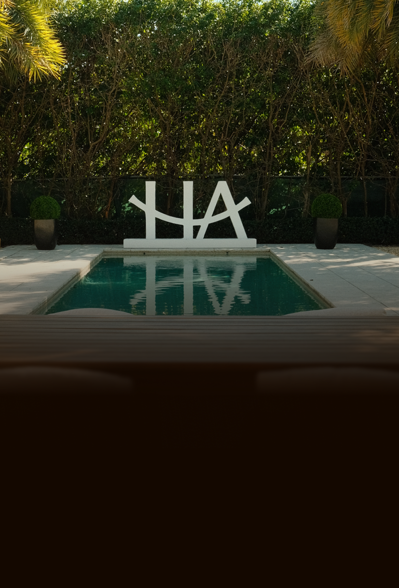 A white abstract sculpture sits at the end of a rectangular swimming pool in front of a tall hedge.