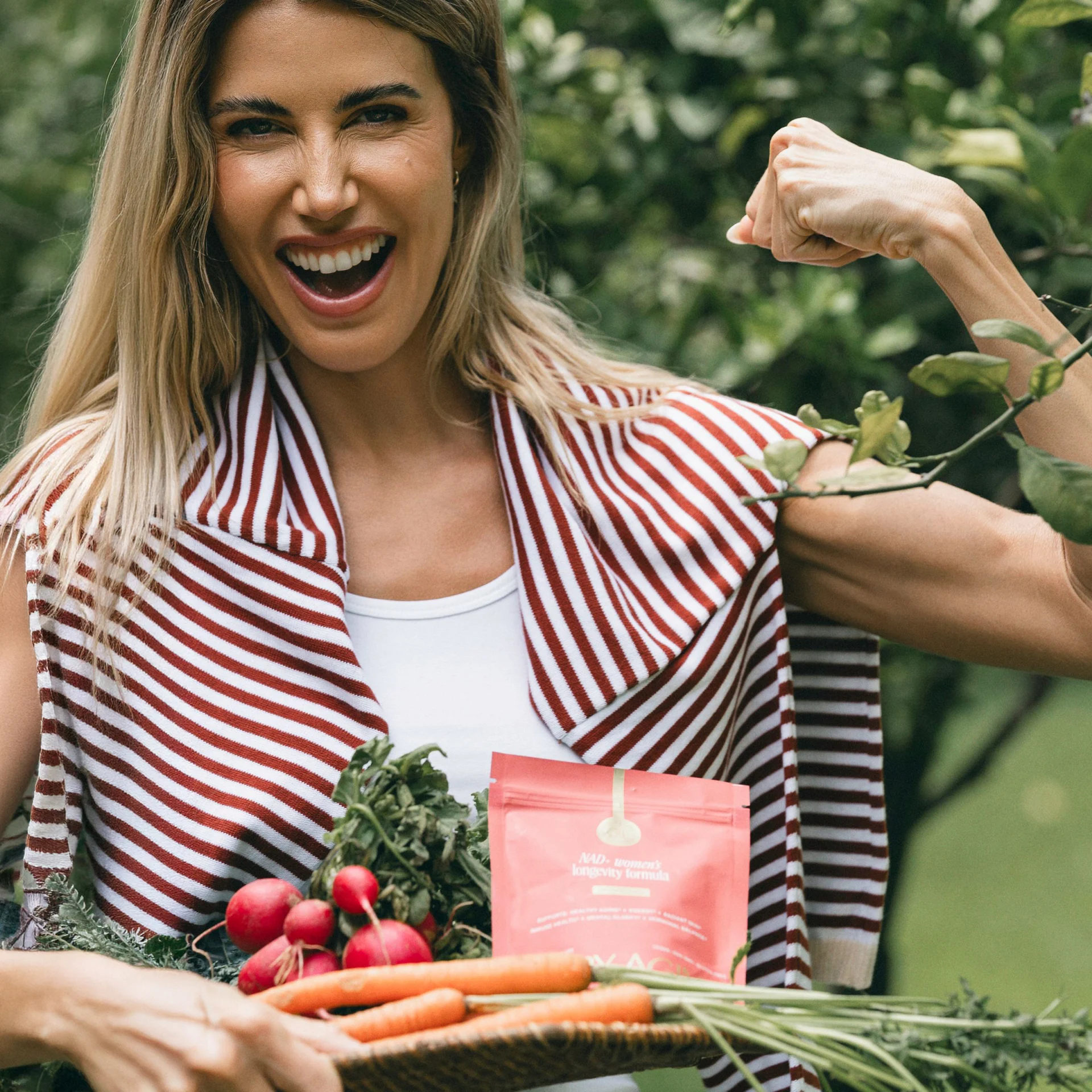 Person smiling, holding a basket with carrots and radishes, wearing a striped scarf.