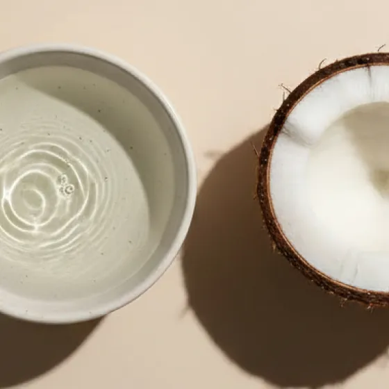 An overhead shot of a halved coconut sitting next to a white bowl of water on a tan surface.