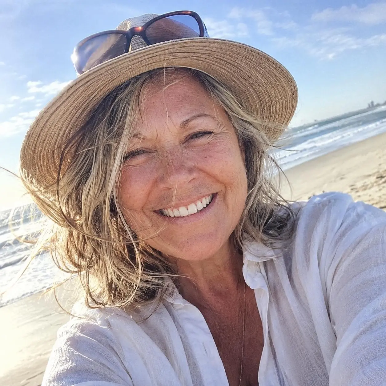 A smiling woman wearing a straw hat and a white shirt takes a selfie on a sunny beach.