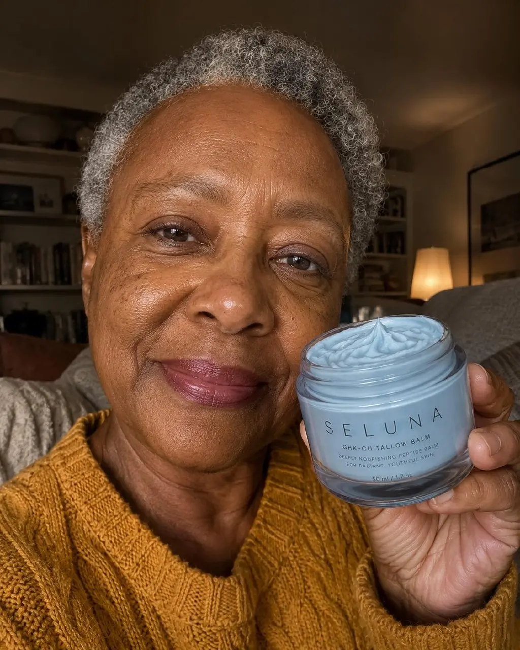 A woman with short gray hair smiles while holding up an open jar of light blue cream.