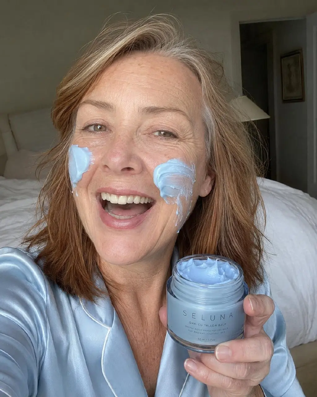 A smiling woman in blue pajamas with light blue cream on her face holds a jar of the product.