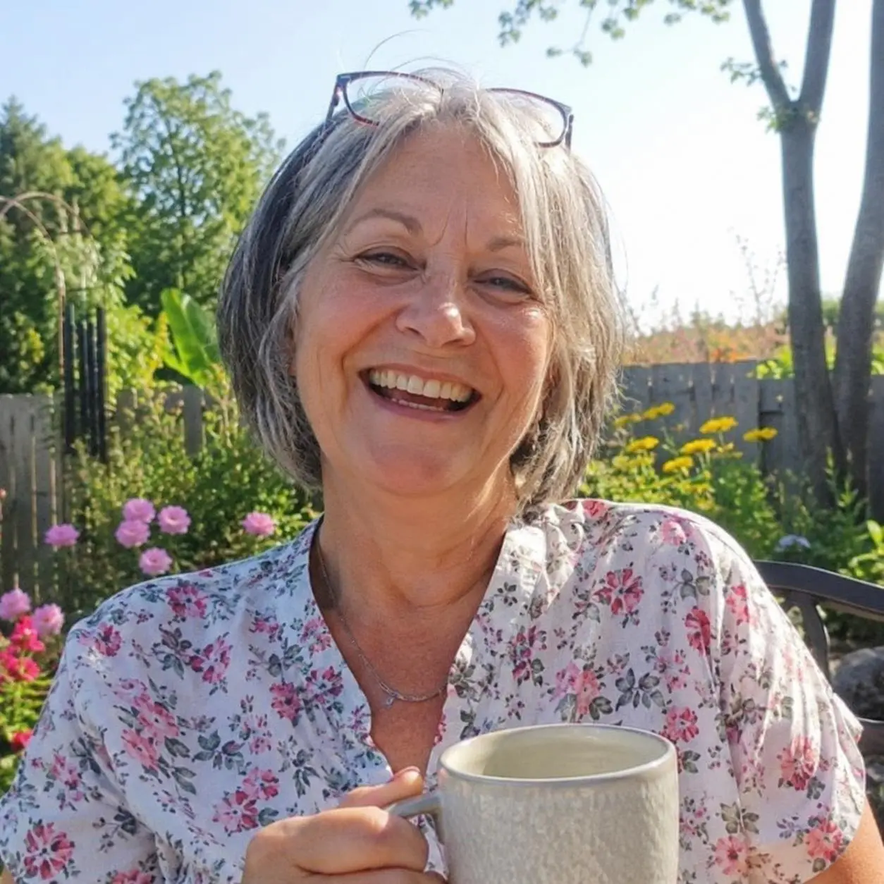 A smiling woman with grey hair and glasses on her head holds a mug in a garden.