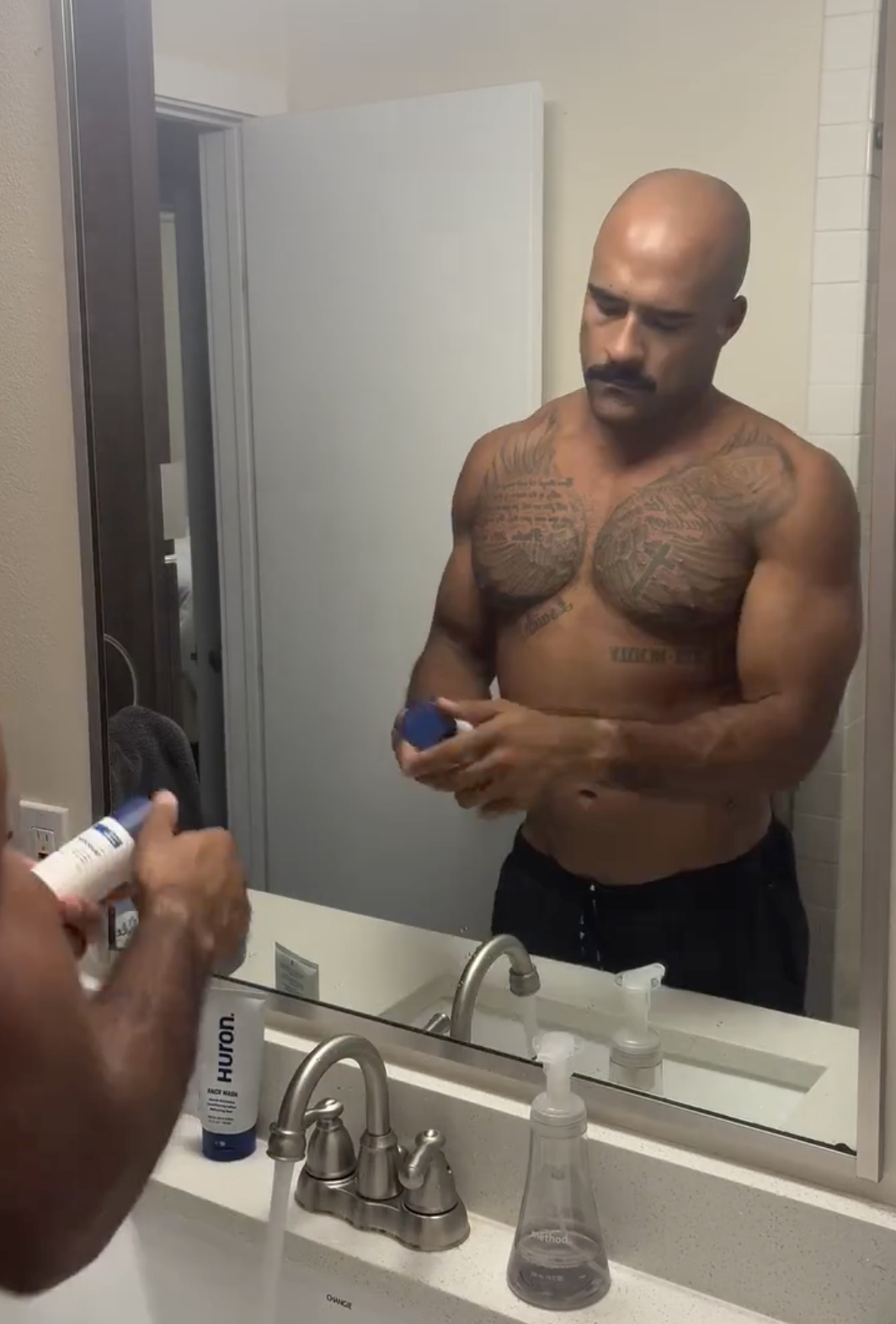 Shirtless man with tattoos holding grooming products in front of mirror.