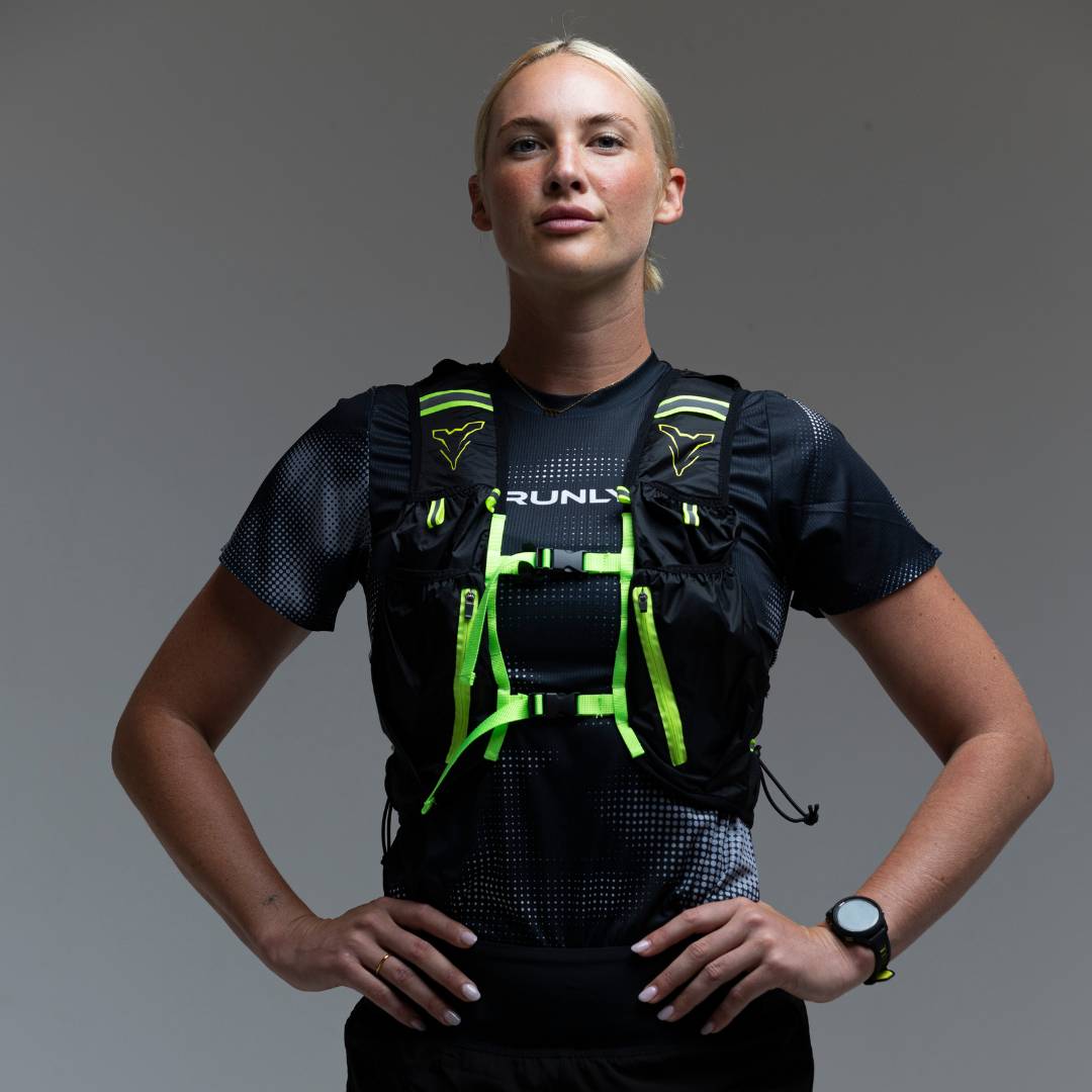 A female runner poses with her hands on her hips, wearing a black and neon green hydration vest.