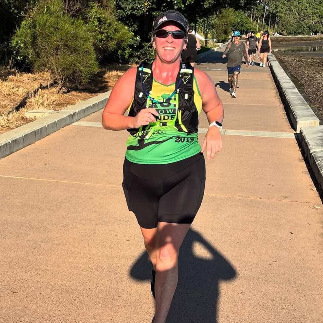 Smiling person running along a paved path surrounded by trees and a few people in the background.