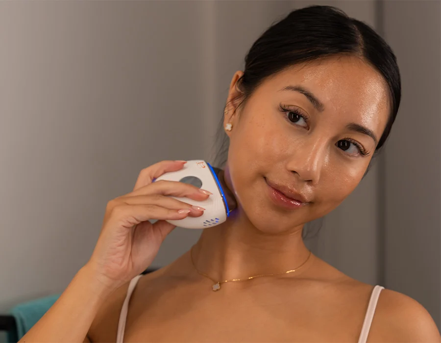 A woman with her hair pulled back uses a handheld beauty device with a blue light on her neck.