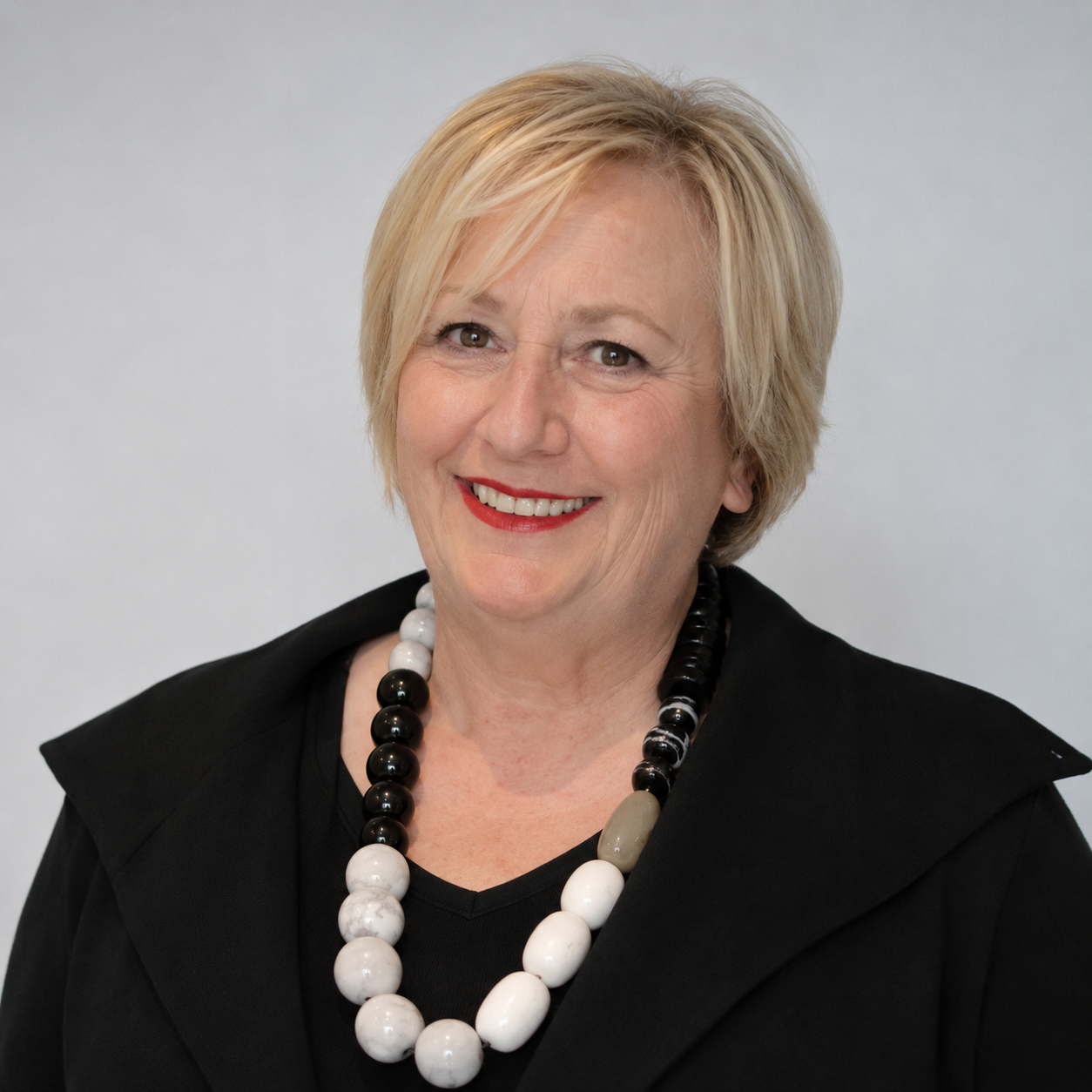 A headshot of a smiling woman with short blonde hair wearing a black jacket and beaded necklace.