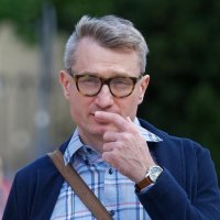 Man with glasses and plaid shirt touching his face outdoors.