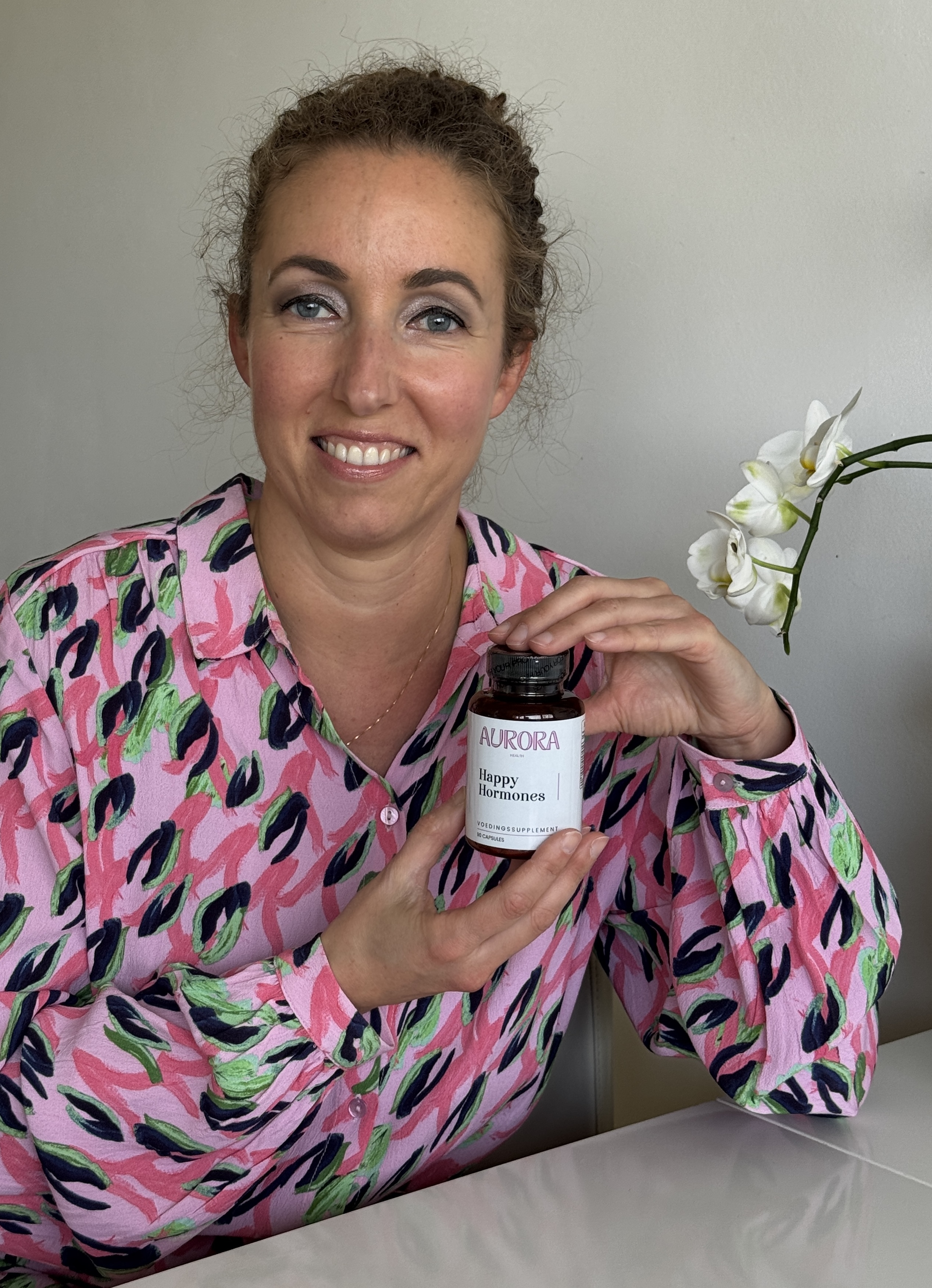 Person holding a bottle labeled 'Aurora Happy Hormones' with a white orchid in the background.