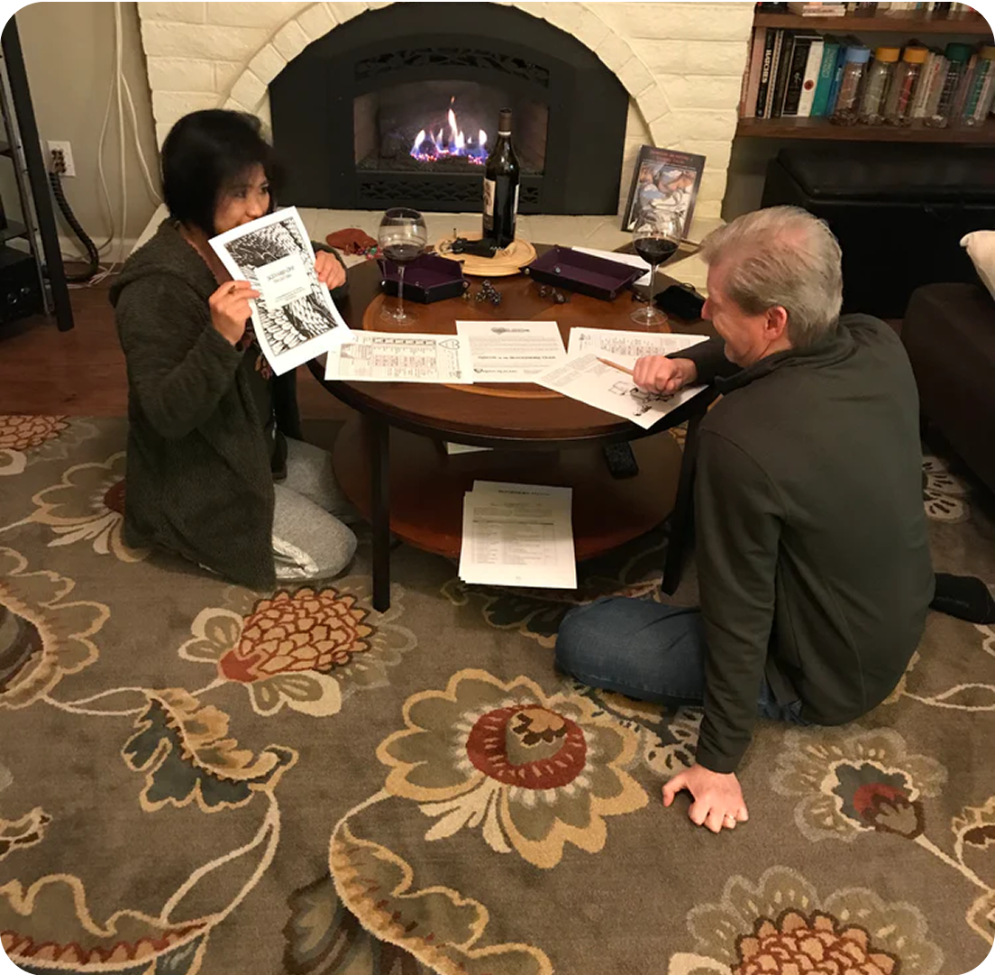 Two people sitting by a coffee table with papers and wine glasses, near a fireplace.