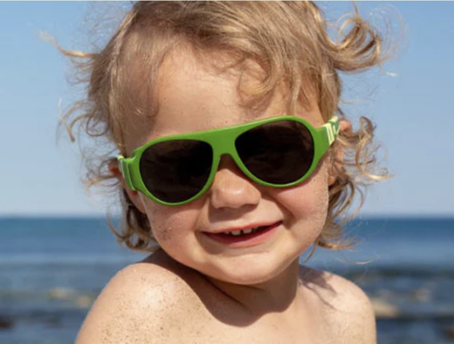 Child with curly hair wearing green sunglasses at the beach.
