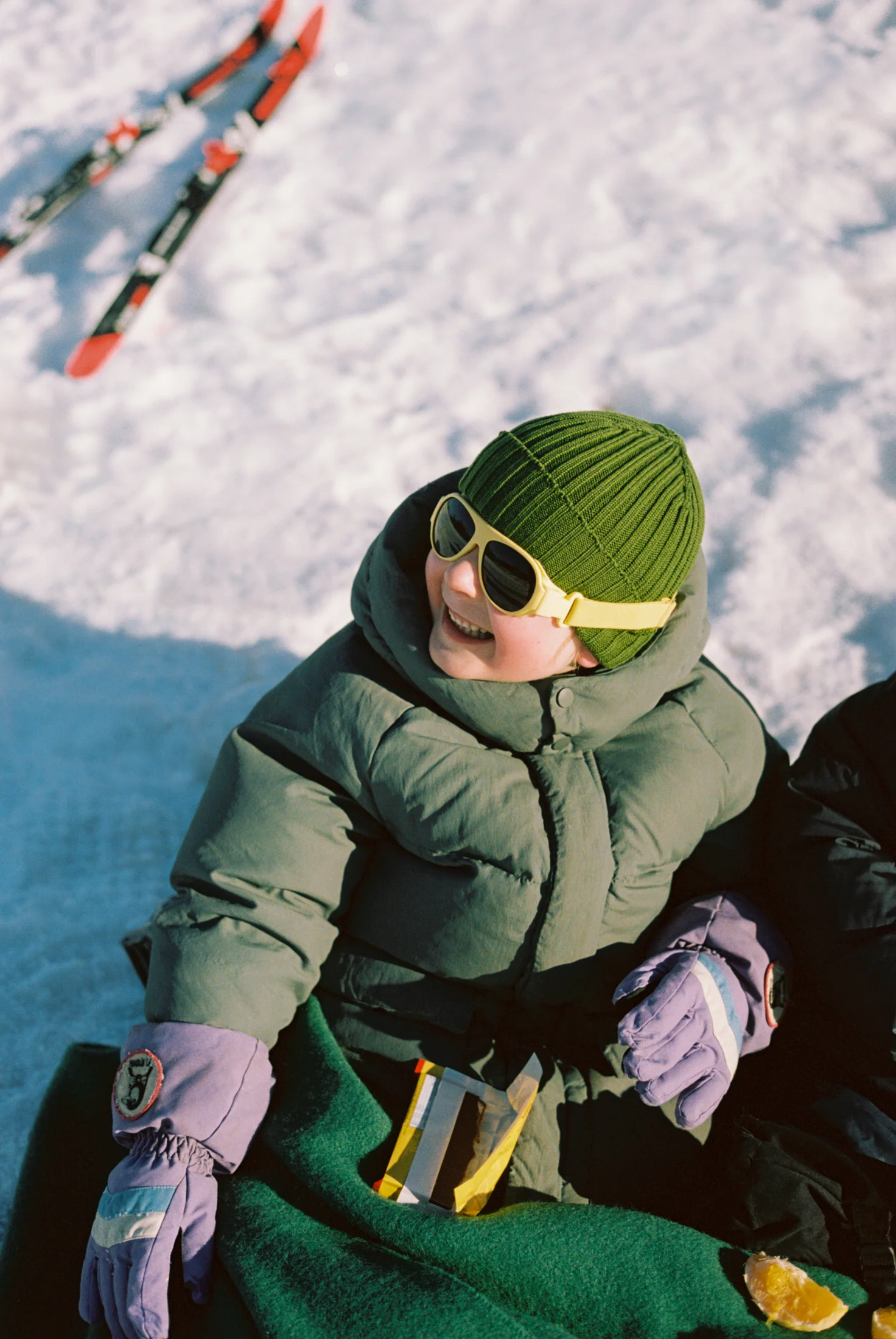 Child in winter clothing smiling on snow with skis in background.