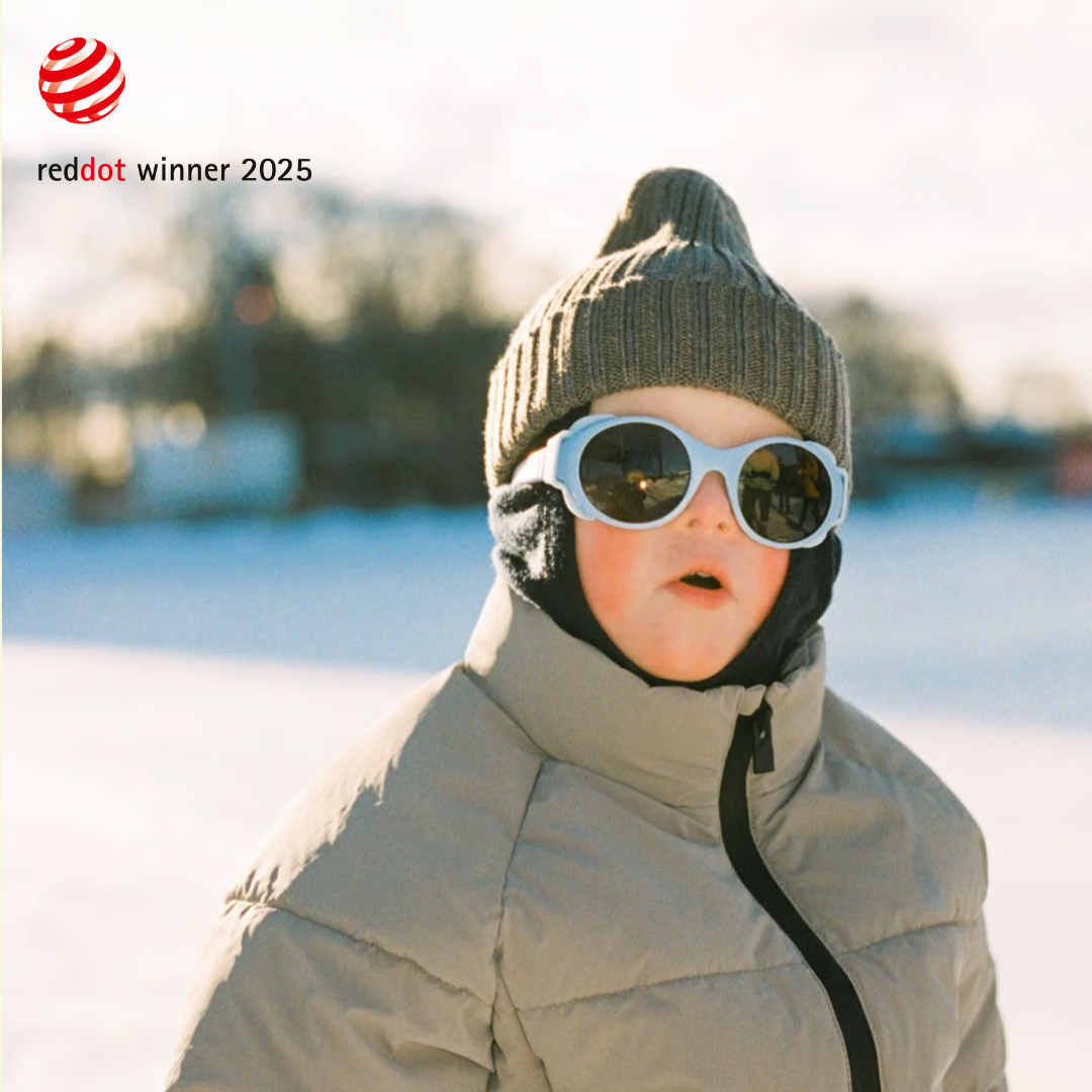 Child in winter clothing and sunglasses, standing outdoors in a snowy landscape.
