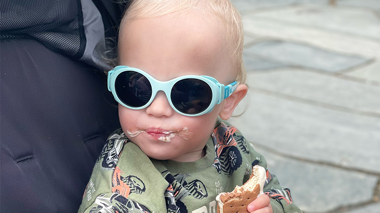 Child wearing sunglasses eating an ice cream sandwich.
