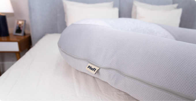 A close-up of a grey Plufl body pillow with a textured cover resting on a bed.