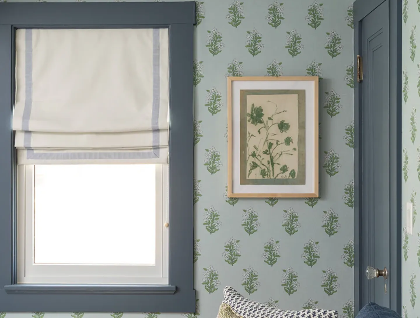 A window with a cream roman shade next to a wall with floral wallpaper and a framed botanical print.