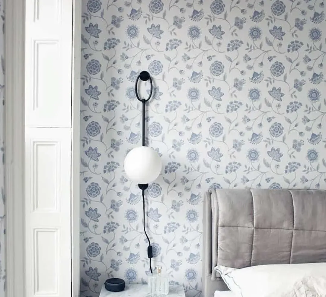 A modern globe lamp hangs on a wall with blue floral wallpaper next to a grey headboard.