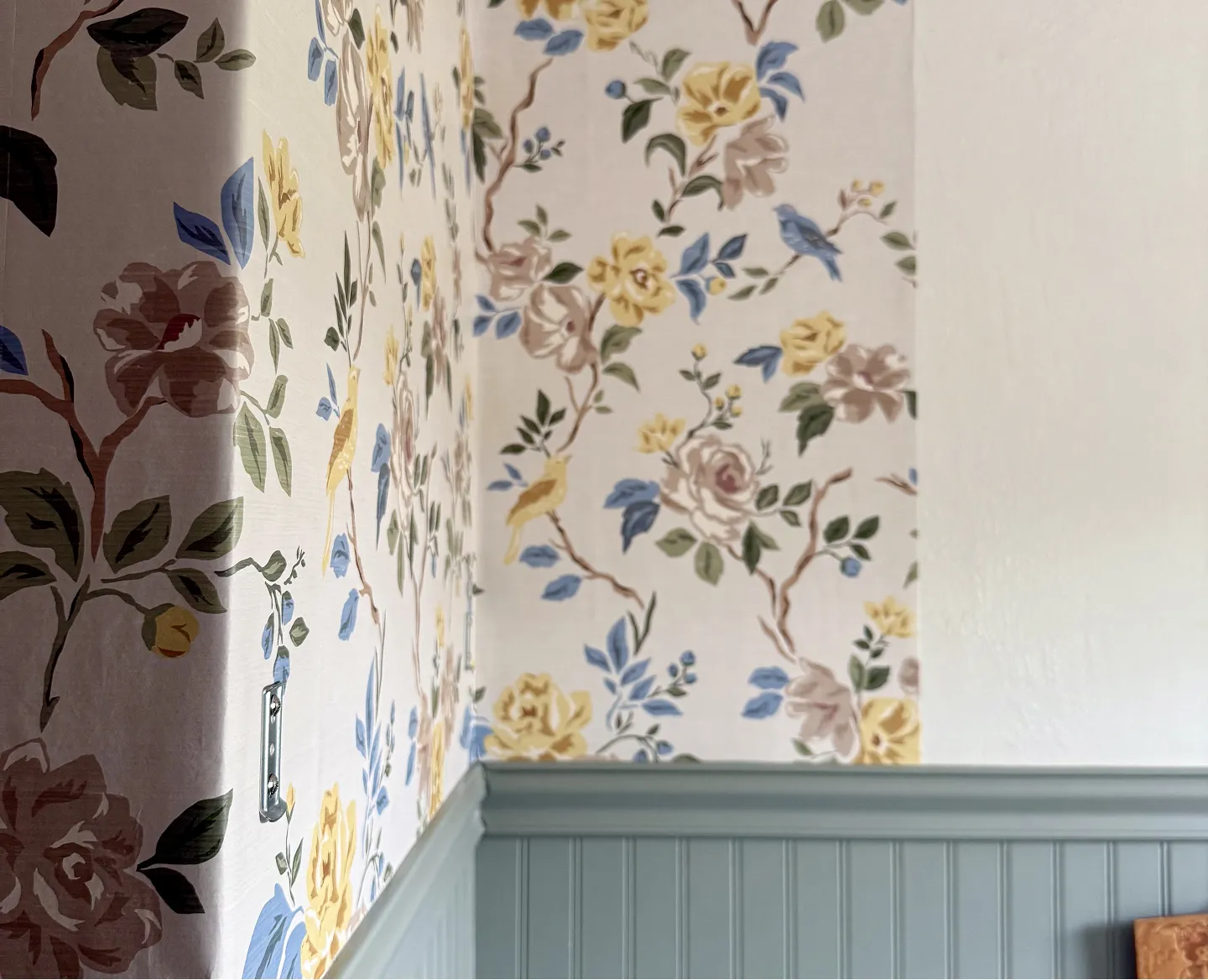 A room corner with floral and bird patterned wallpaper above blue-gray beadboard wainscoting.