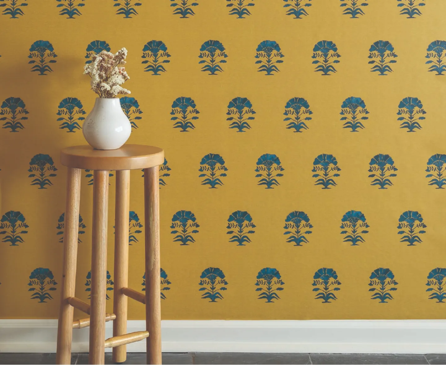A white vase with dried flowers on a wooden stool against a yellow wall with blue floral patterns.