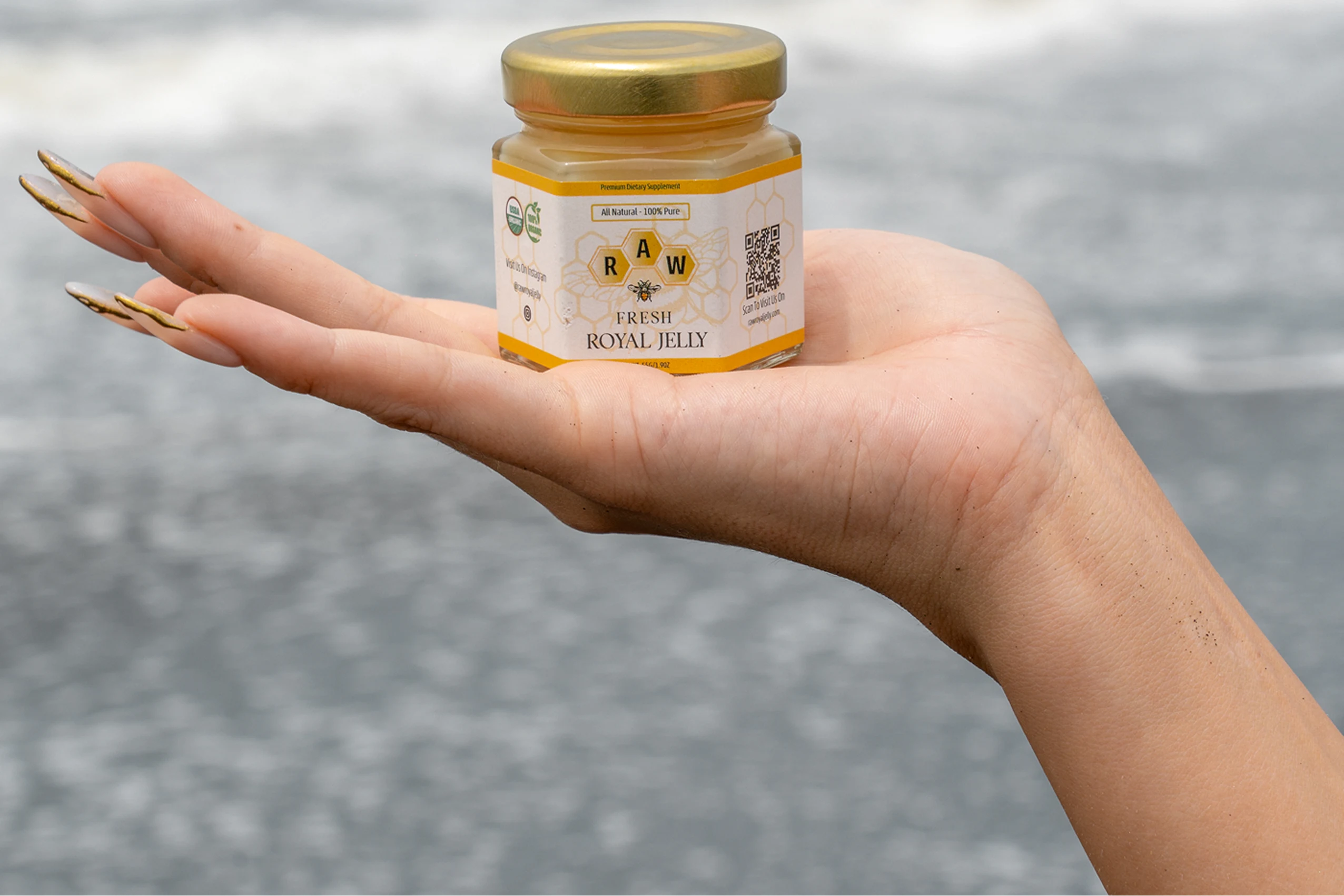 A person's hand holding a small jar of Raw Fresh Royal Jelly in front of water.