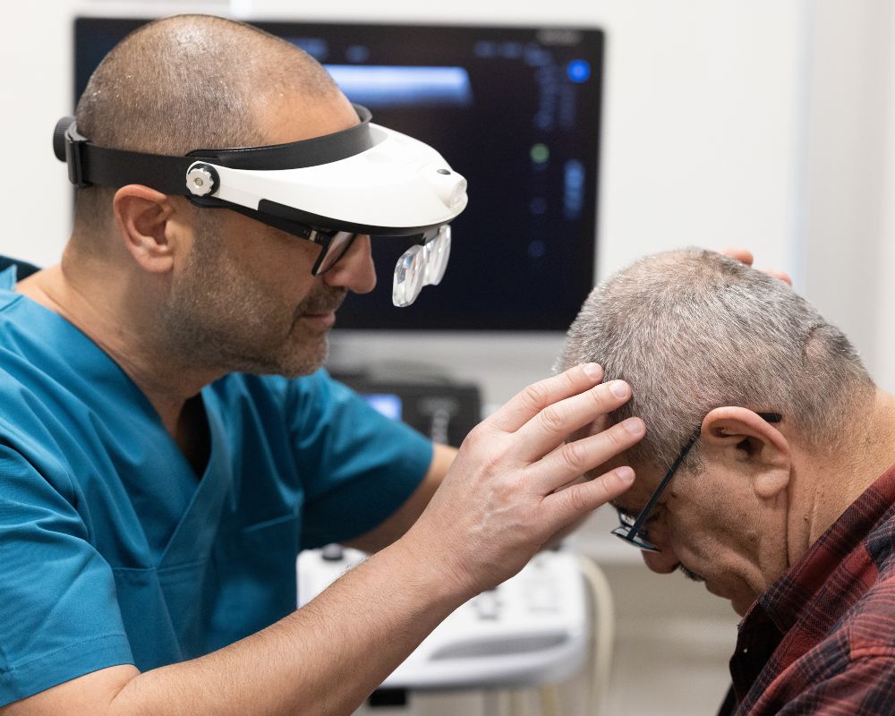 A medical professional wearing a head-mounted magnifier examines a patient's scalp in a clinical setting.