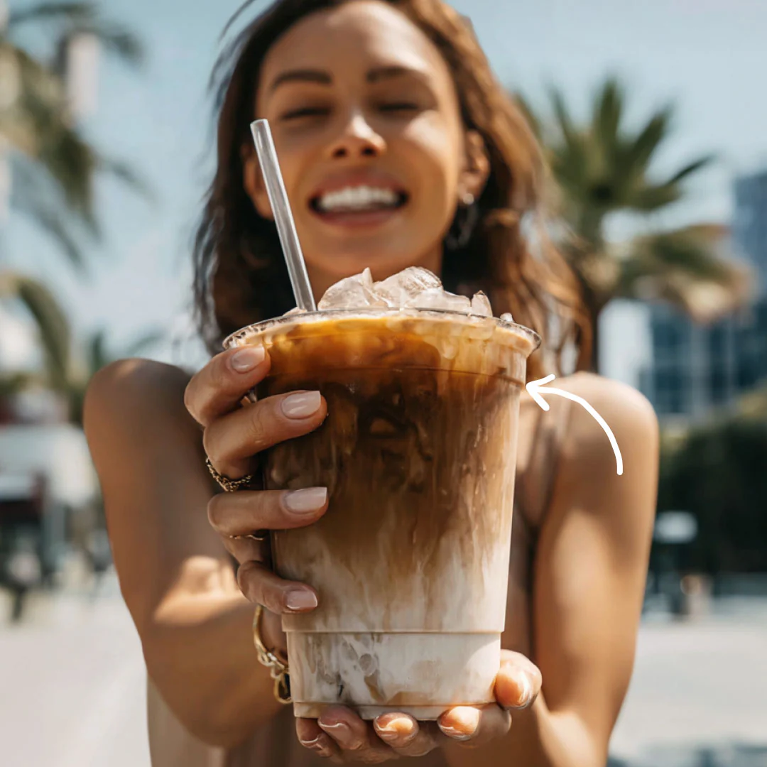 A smiling woman holds a large cup of layered iced coffee towards the camera on a sunny day.