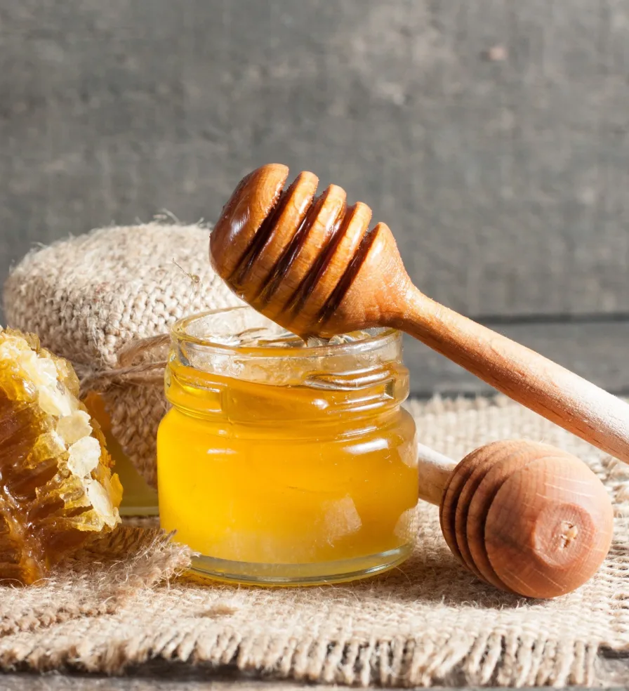 Jar of honey with dipper and honeycomb on a woven mat.