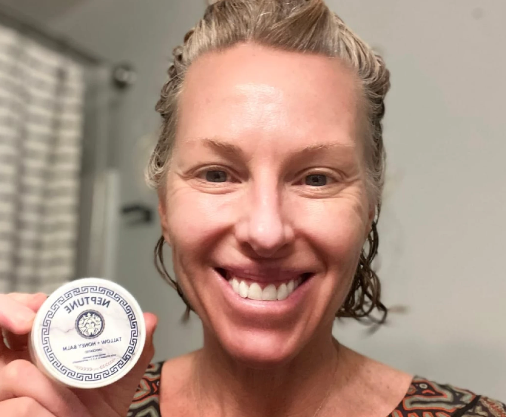 A smiling woman with damp blonde hair holds up a small, round container of Neptune skin balm.