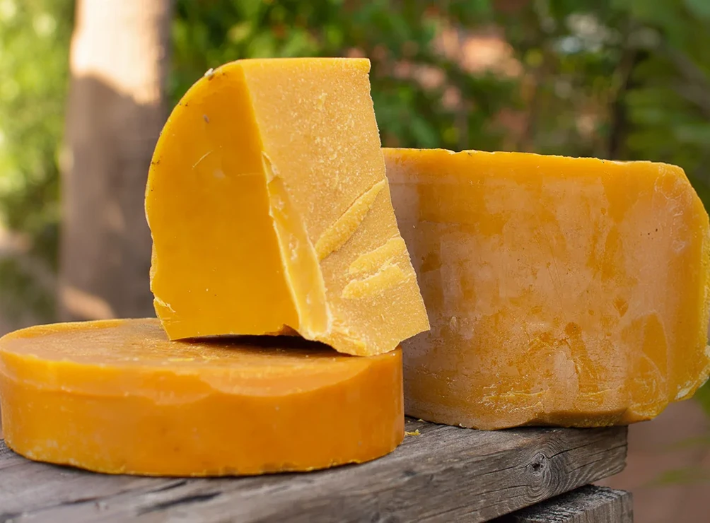 Three large blocks of yellow beeswax sitting on a rustic wooden plank outdoors.