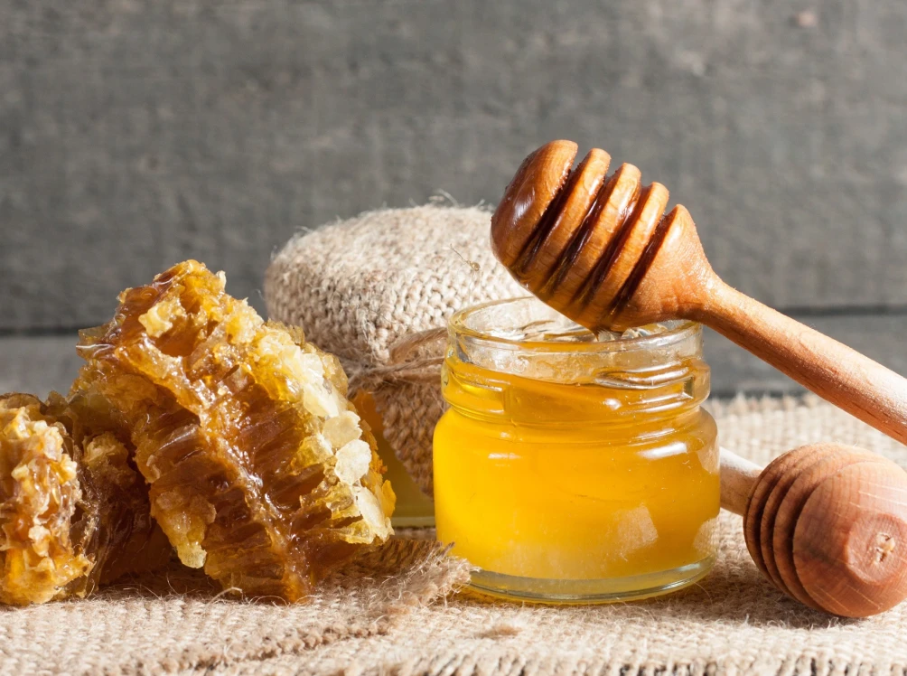 A jar of honey with a wooden honey dipper resting on it, next to a piece of honeycomb.